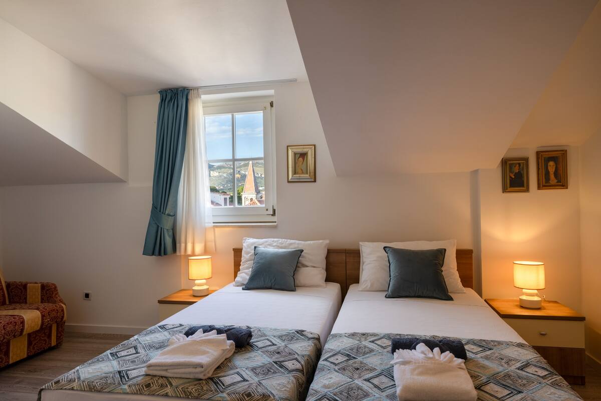 A hotel room with two beds, a window with curtains, a nightstand with a lamp, and a painting on the wall.
