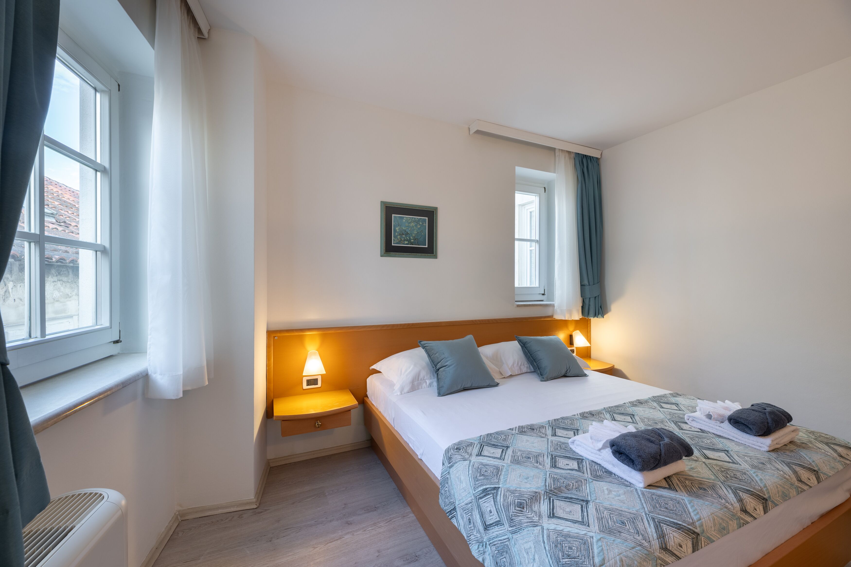 A neatly made bed with a patterned bedspread, two pillows, and folded towels on the side. There is a bedside lamp, a framed picture on the wall, and a window with curtains.
