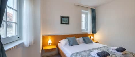 Una cama bien tendida con una colcha estampada, dos almohadas y toallas dobladas al lado. Hay una lámpara de noche, un cuadro enmarcado en la pared y una ventana con cortinas.