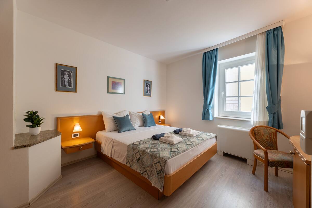 A hotel room with a bed, bedside tables, a chair, a desk, a window with curtains, and wall art.