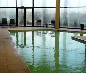 An indoor swimming pool with clear glass walls, a tiled floor, and several chairs around the pool.