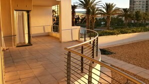 Ein moderner Hotelbalkon mit gebogener Geländer, gefliestem Boden und Blick auf Palmen sowie ein Gebäude im Hintergrund.