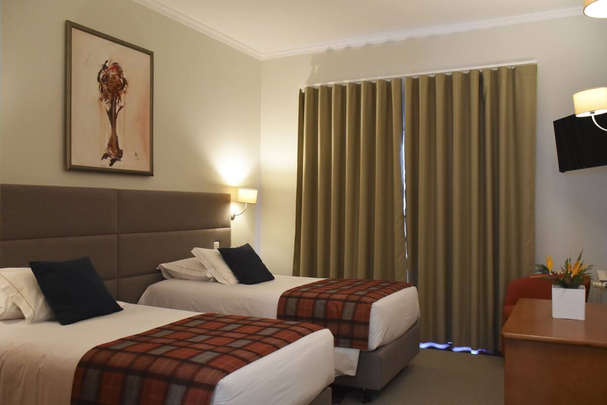 A hotel room with two beds, a headboard, a desk with a chair, a television, and a window with curtains.