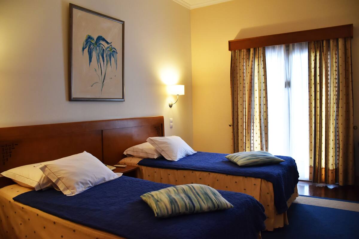 A hotel room with two beds, a nightstand, a lamp, a painting, and a window with curtains.