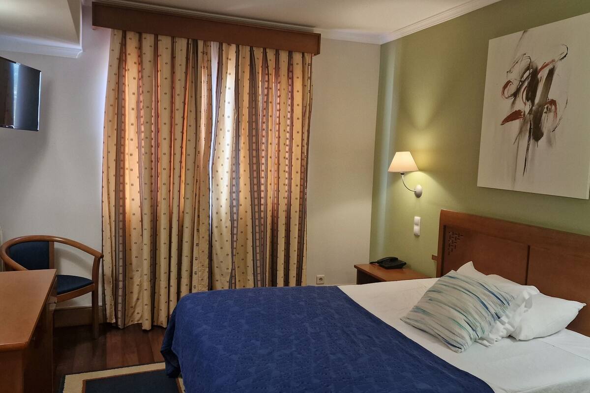 A hotel room with a bed, a desk, a chair, a lamp, and a painting on the wall.