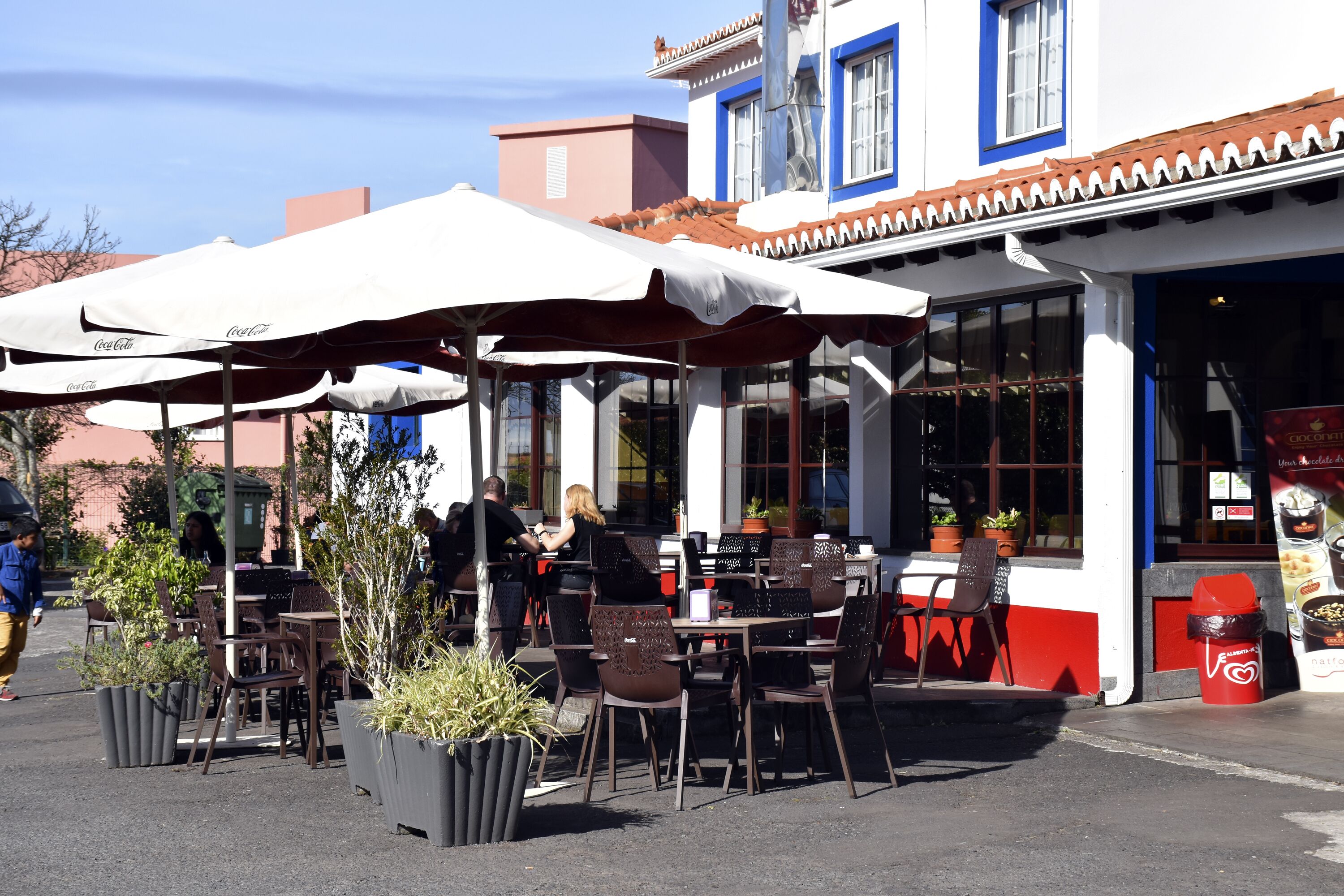 An outdoor cafe with tables and chairs under umbrellas, potted plants, and a building with large windows in the background.