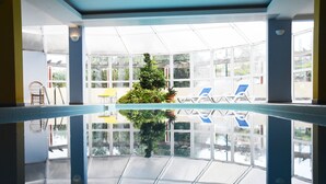 Un área de piscina interior amplia con paredes de vidrio transparente, un árbol de Navidad en el centro y varias sillas de descanso.