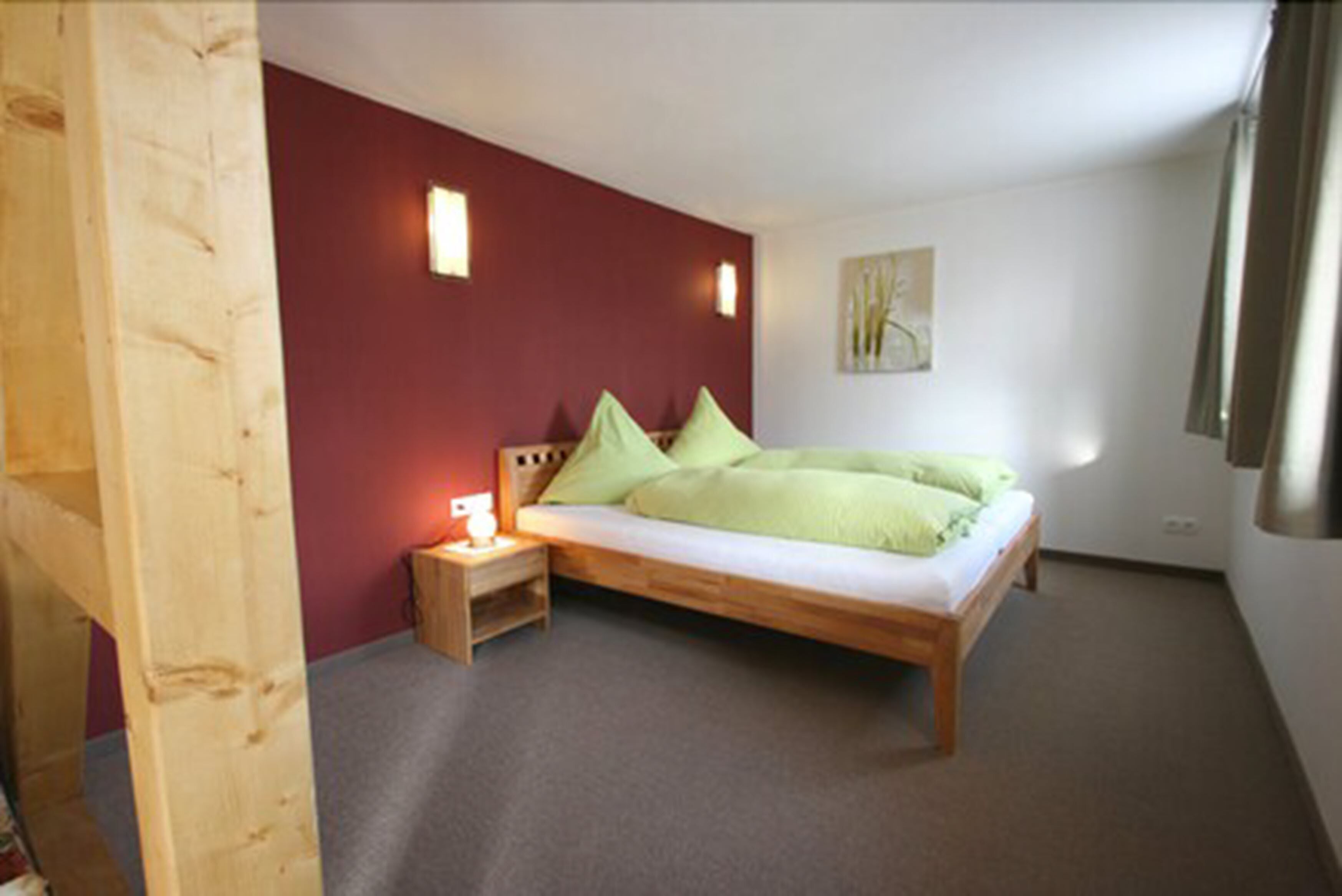 A bedroom with a wooden bed, green pillows, a nightstand with a lamp, and a red accent wall.