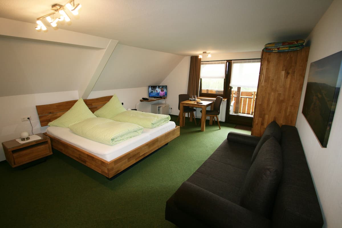 Quarto com cama, mesa de cabeceira de madeira, escrivaninha com cadeira, sofá marrom e televisão em móvel.