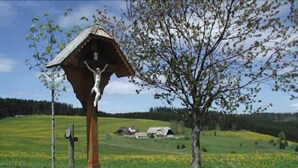 Ländliche Landschaft mit einem hölzernen Kruzifix auf einem Pfosten, einem Baum und einem Haus im Hintergrund.