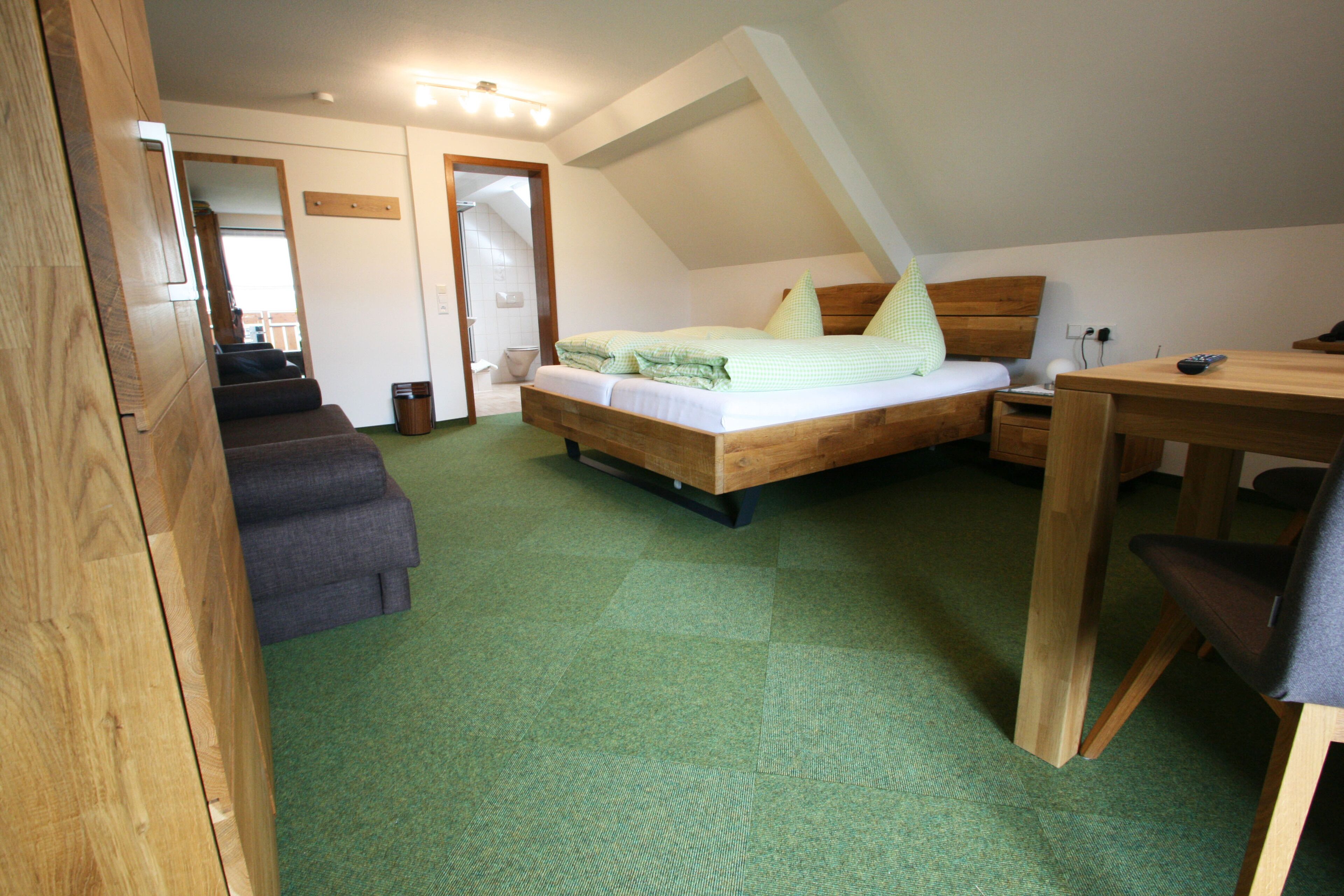 A bedroom with a wooden bed, a desk, and a chair. There is a bathroom visible through an open door.