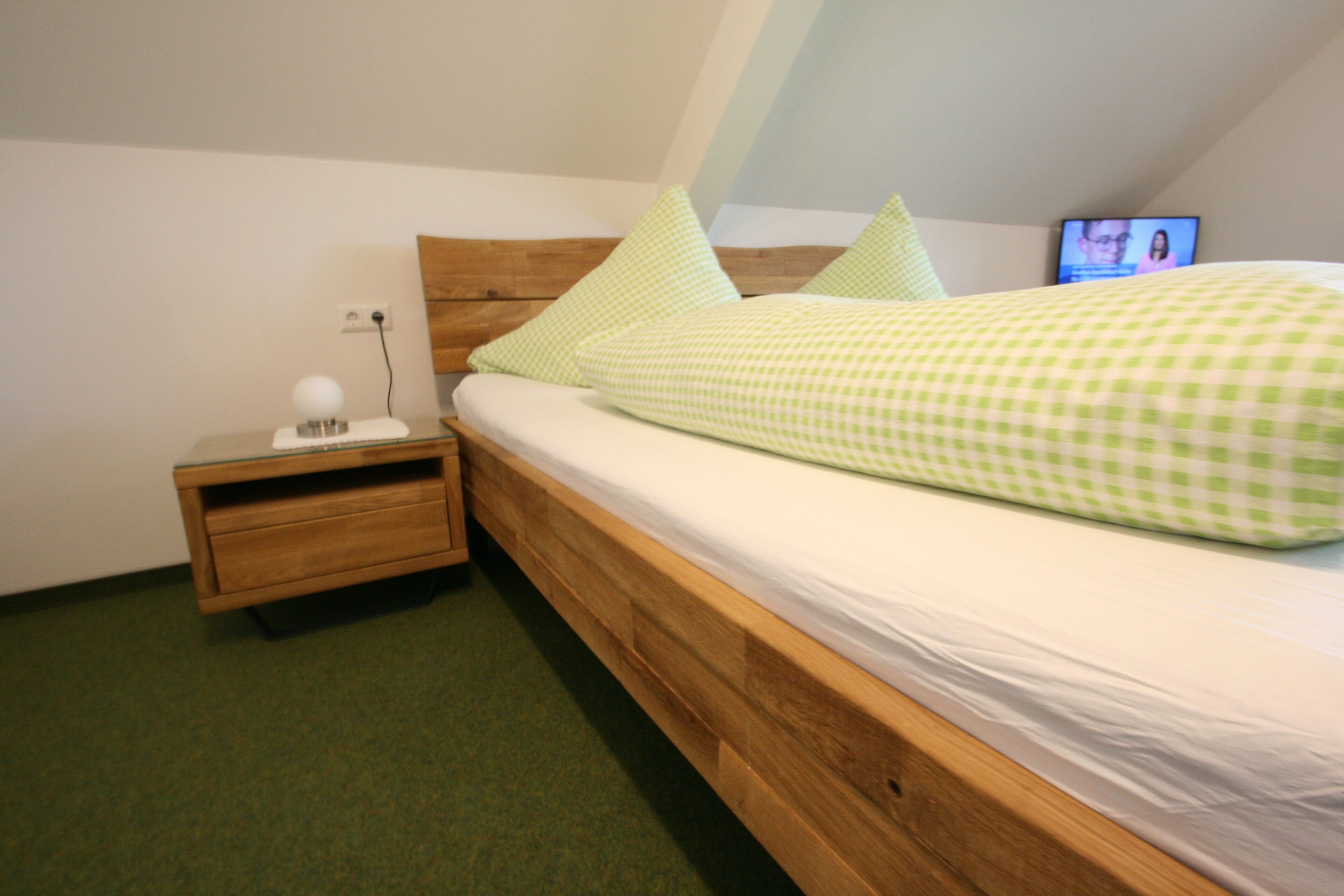 A single bed with a wooden frame and a green checkered bedspread, a nightstand with a lamp, and a television on the wall.