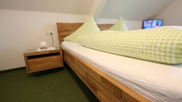 A single bed with a wooden frame and a green checkered bedspread, a nightstand with a lamp, and a television on the wall.