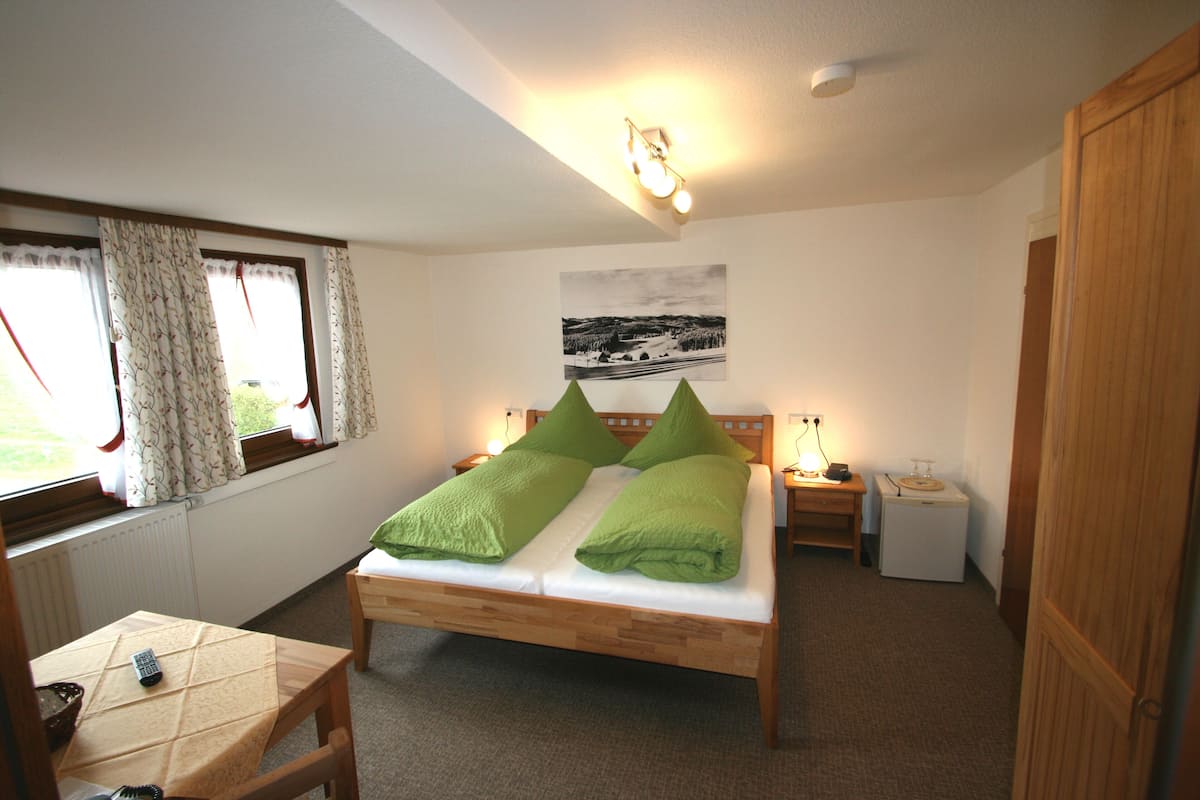 Quarto com cama de madeira, duas mesas de cabeceira, um criado-mudo, janela com cortinas, radiador e um quadro na parede.