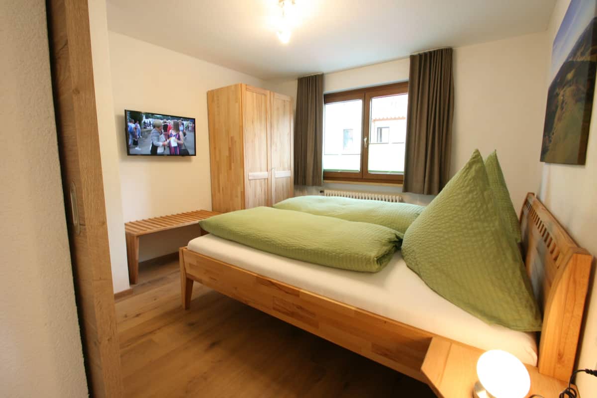 Um quarto com uma cama de madeira, um banquinho, um guarda-roupa e uma TV na parede.