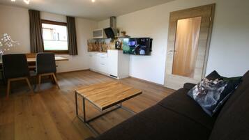 Um espaço de estar compacto com piso de madeira, um sofá, uma mesa de jantar com cadeiras, uma cozinha com micro-ondas e uma TV, e uma porta.