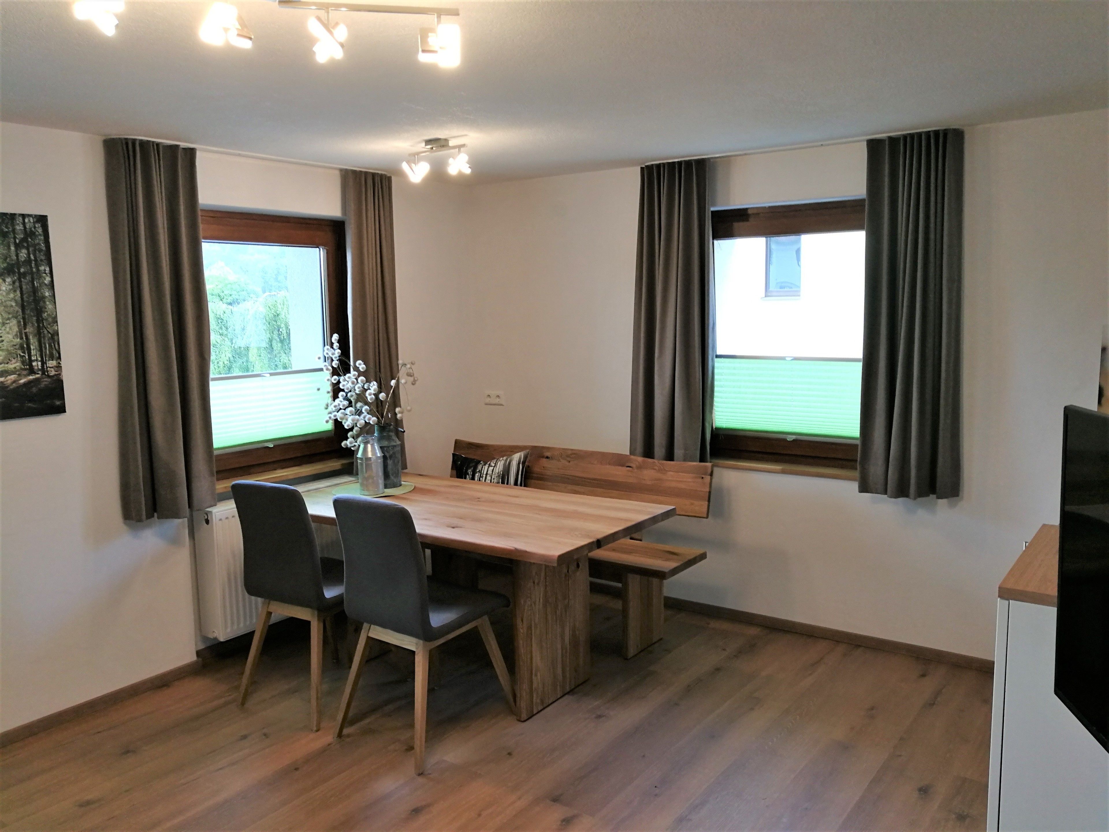 Un comedor con una mesa de madera, cuatro sillas, un jarrón con flores y dos ventanas con cortinas.