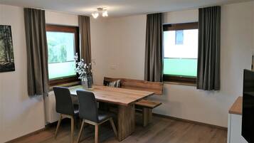 Un comedor con una mesa de madera, cuatro sillas, un jarrón con flores y dos ventanas con cortinas.