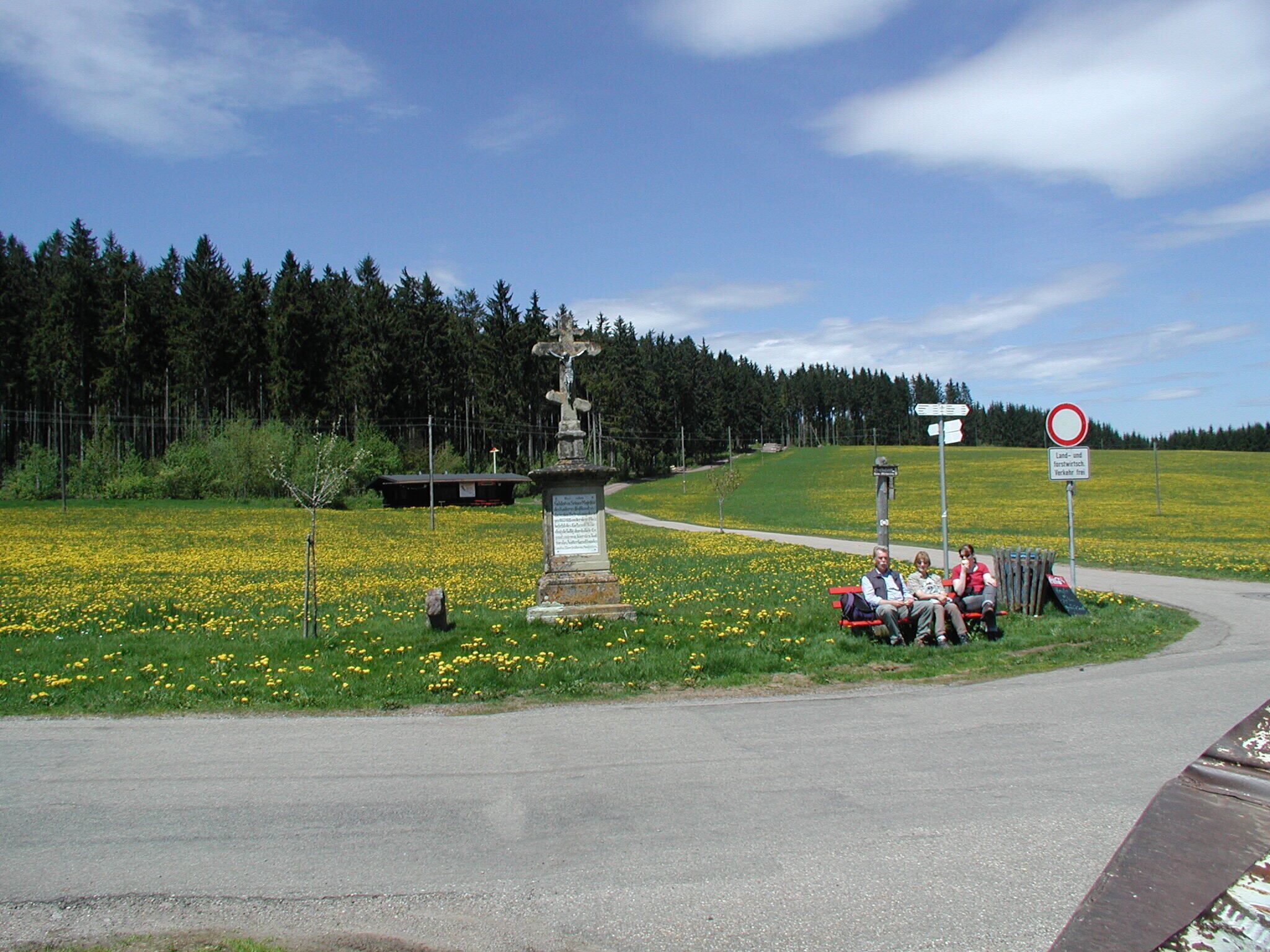 A rural road with a stone monument, a bench, and a signpost in a field of yellow flowers.