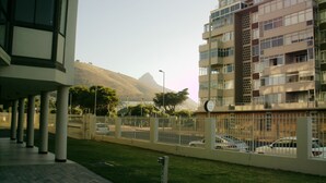 Un moderno edificio de apartamentos con varios balcones, un estacionamiento con coches y una montaña al fondo.