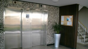Ascensor con pantalla digital, un tablón de anuncios, una planta en maceta y una escalera.