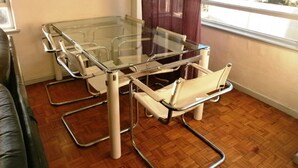 A room with a glass-top table and metal chairs, wooden flooring, a window with a view of buildings, and a brown curtain. - Glenlodge 401 - Apartment (Cape Town)