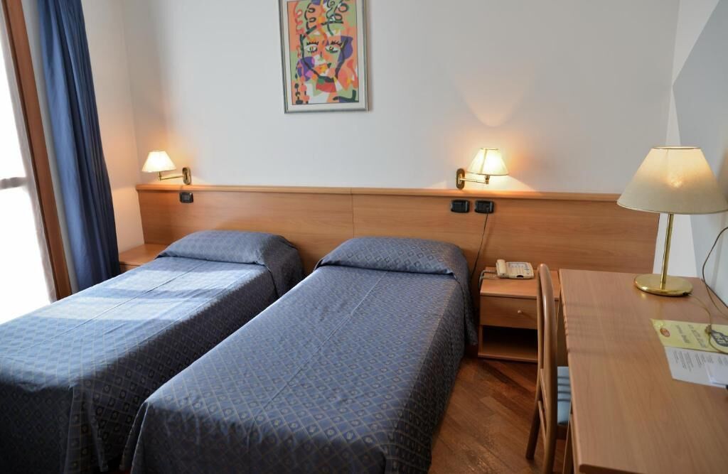 Camera d'albergo con due letti singoli, una testiera in legno, lampade ai comodini, una scrivania con lampada e un quadro appeso al muro.