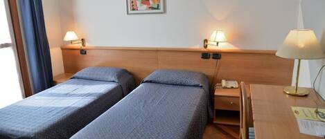 Camera d'albergo con due letti singoli, una testiera in legno, lampade ai comodini, una scrivania con lampada e un quadro appeso al muro.
