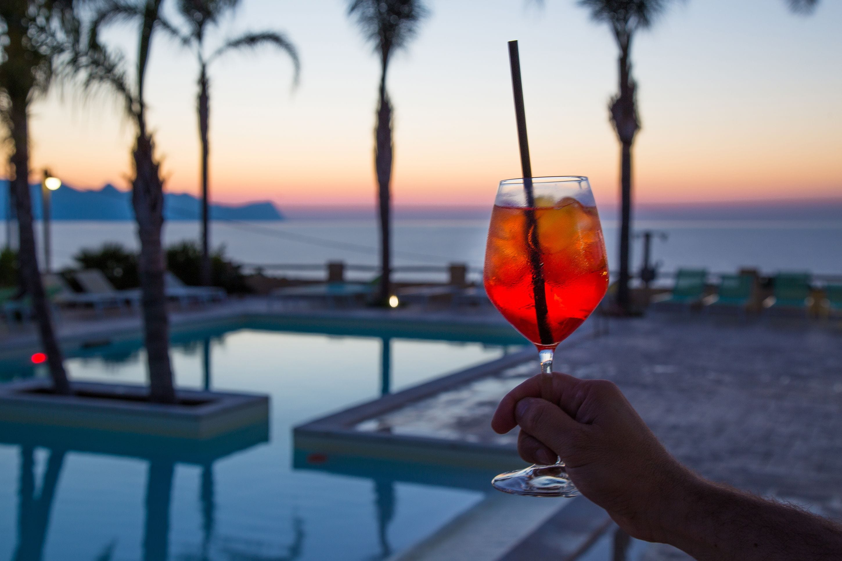 A hand holding a glass with a straw, overlooking a pool with palm trees and a sunset view.