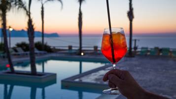A hand holding a glass with a straw, overlooking a pool with palm trees and a sunset view.