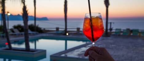 Una mano sosteniendo un vaso con una pajita, con vistas a una piscina, palmeras y una puesta de sol.