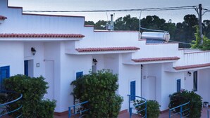 A coastal building with white walls and red-tiled roofs, featuring a staircase and a view of the sea and mountains in the background.