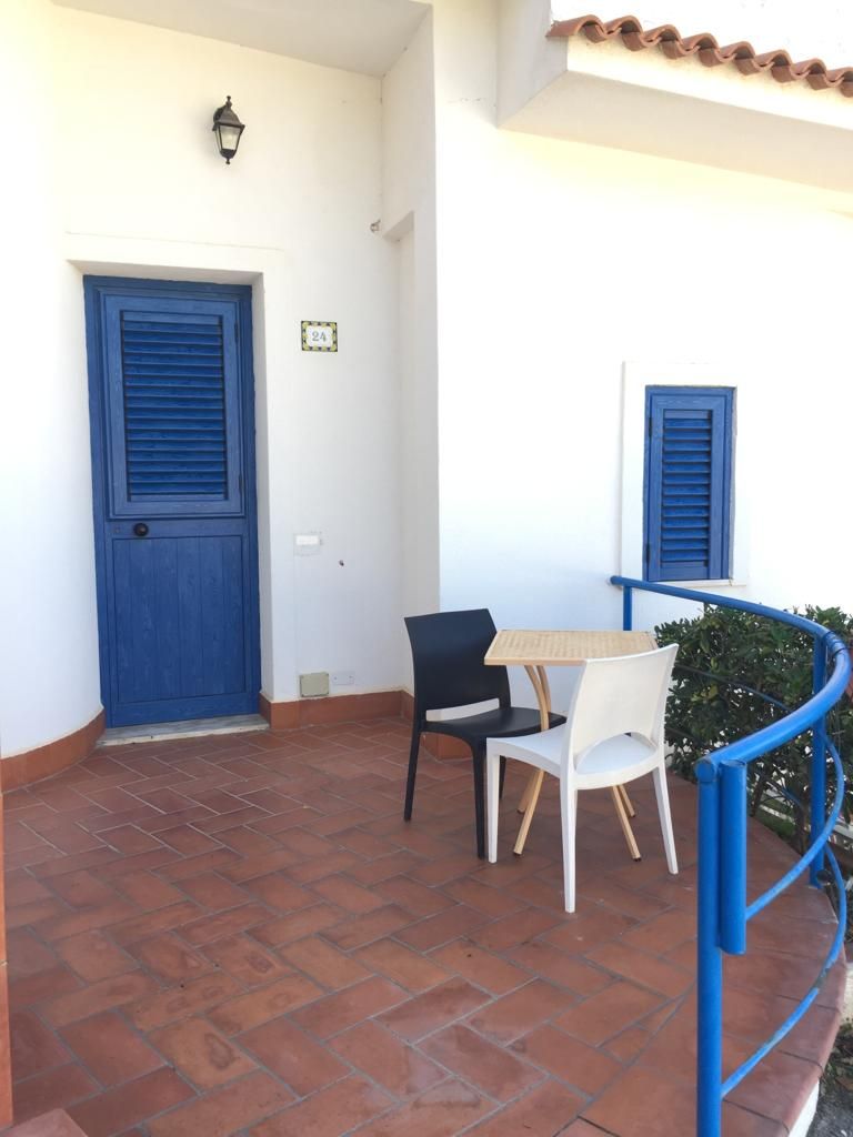 Un balcón con una puerta azul, una mesa con dos sillas y una pequeña planta en maceta.