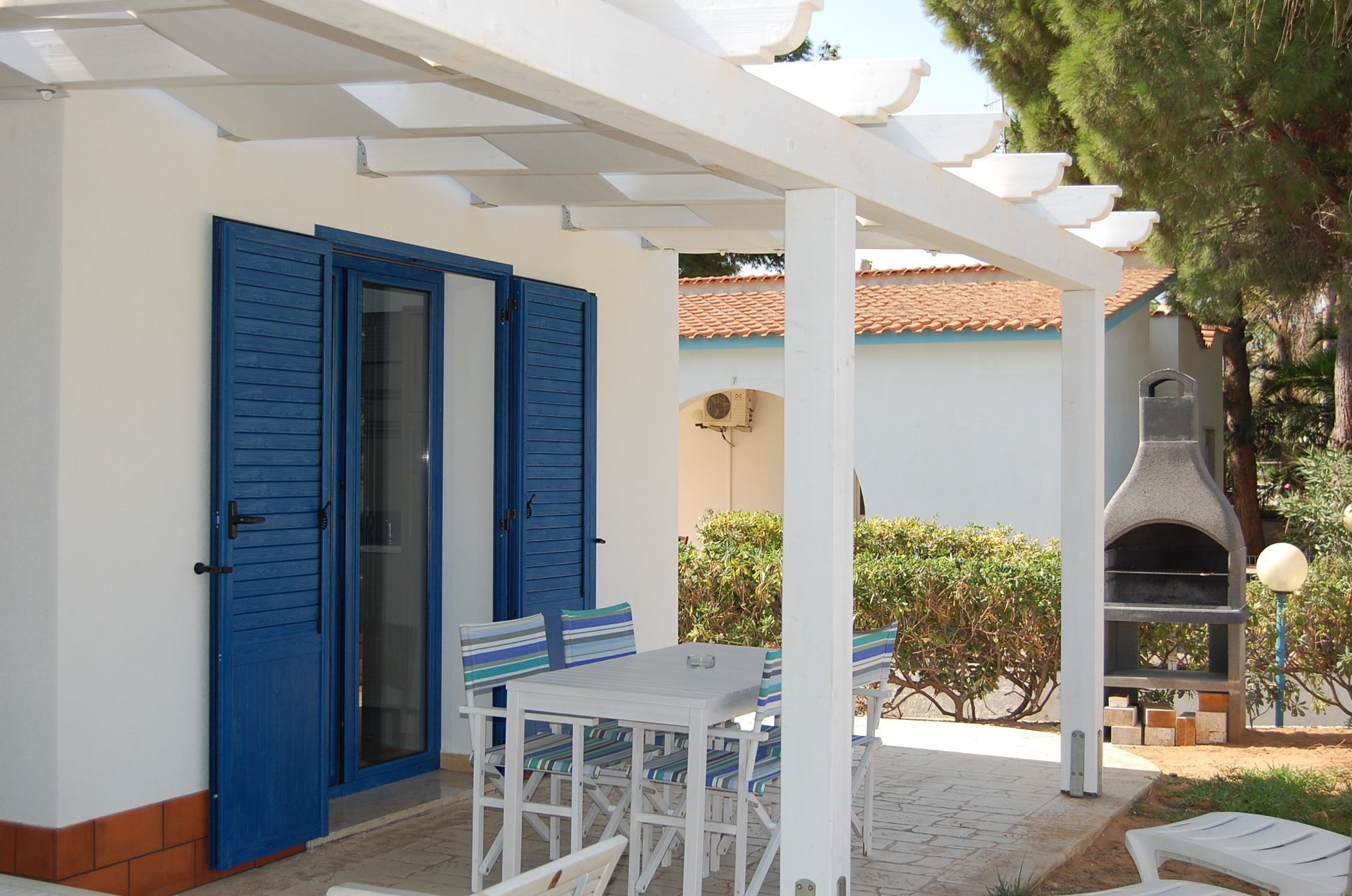 A covered outdoor area with a white table and chairs, a blue door, and a stone-built barbecue grill.