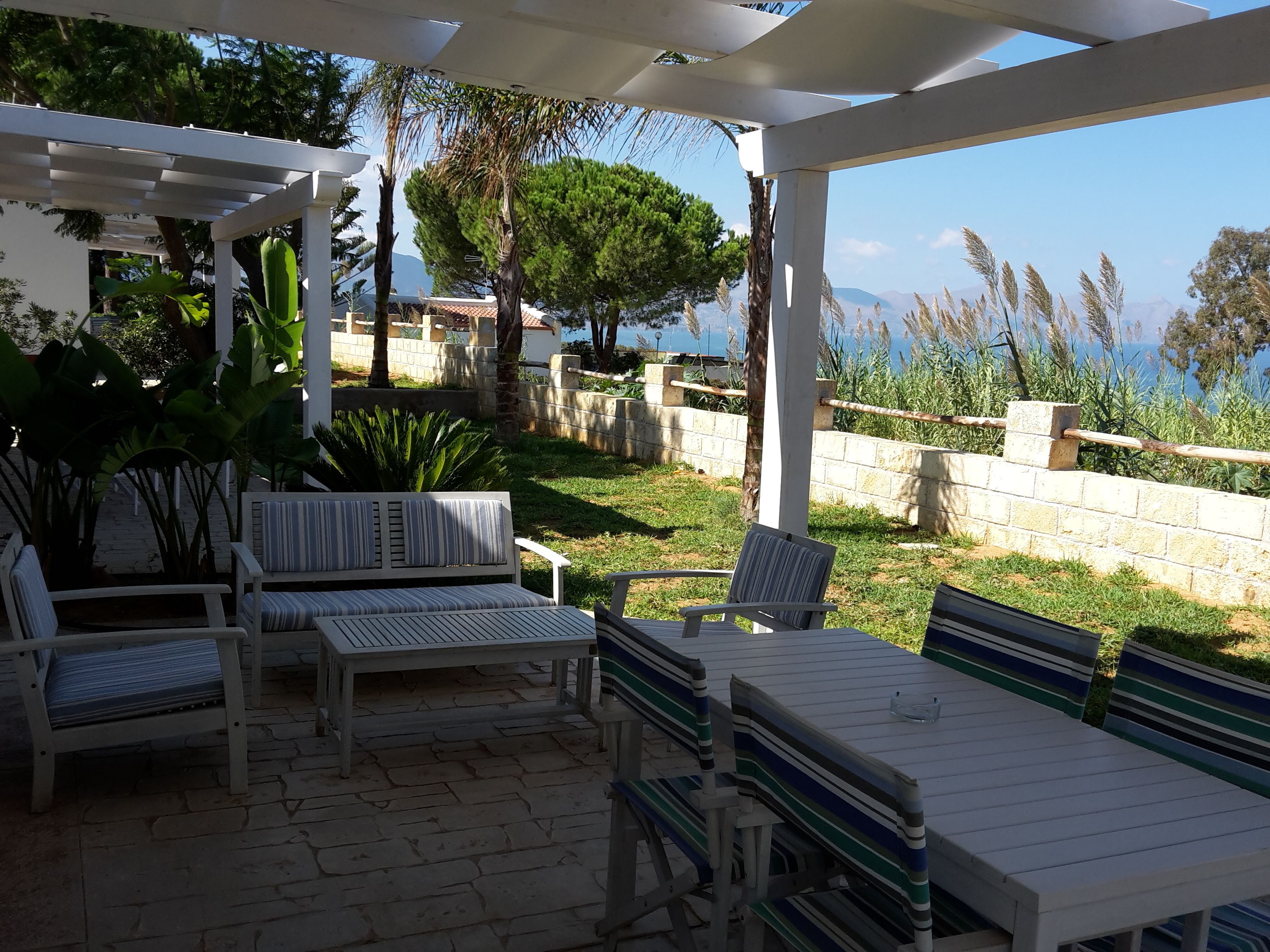 Une terrasse extérieure meublée de mobilier blanc, comprenant des bancs et une table, entourée de verdure et d’un mur en pierre.