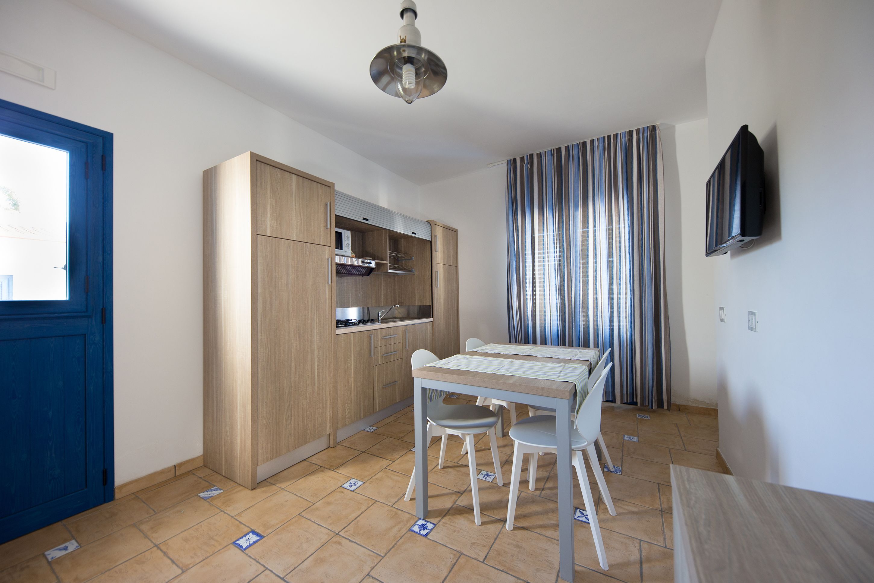 A compact kitchen with wooden cabinets, a dining table with chairs, and a TV mounted on the wall.