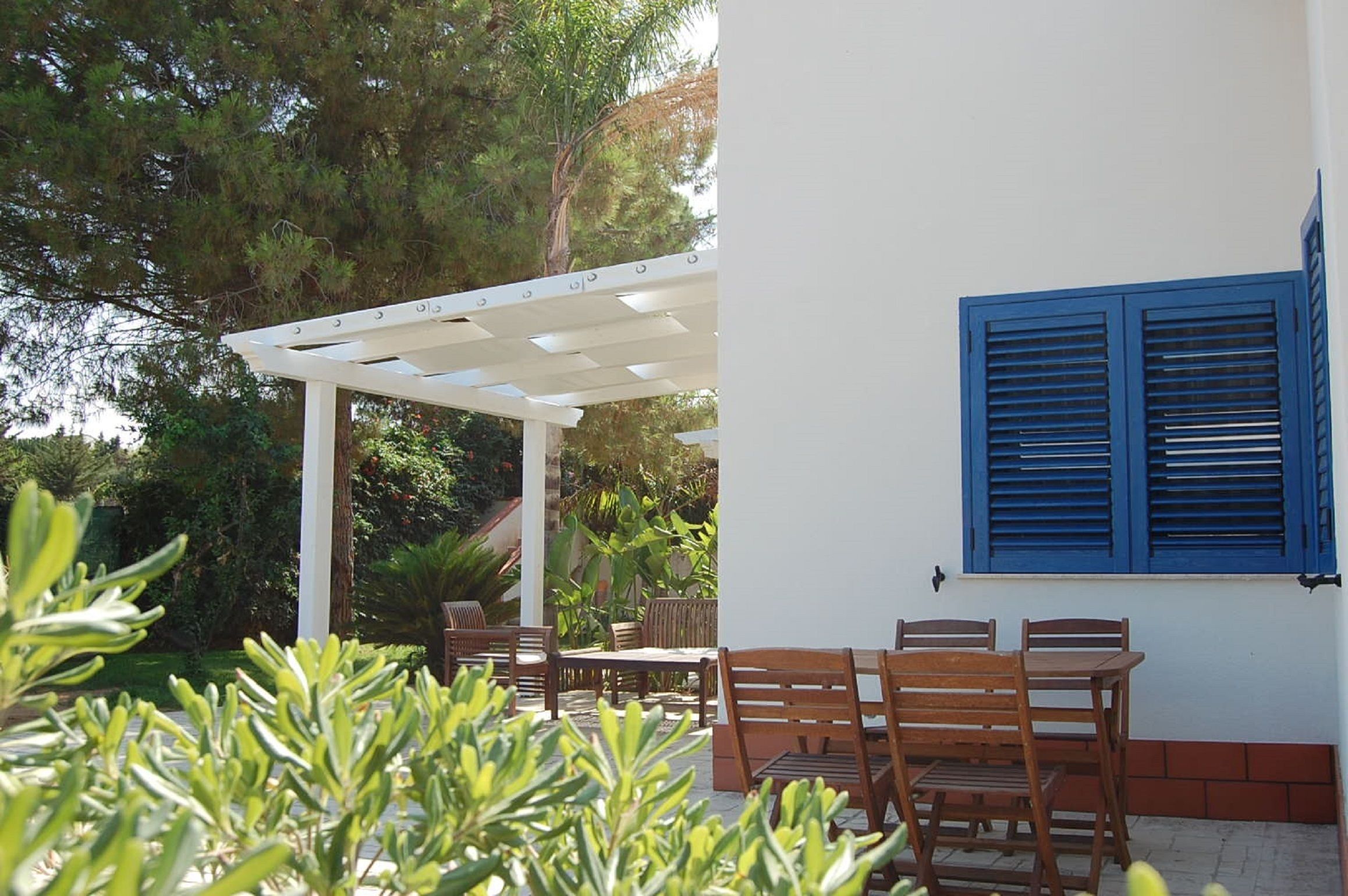A patio with wooden furniture and a white pergola, surrounded by greenery.