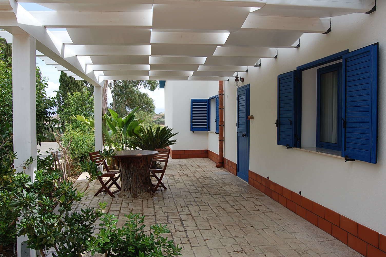 A covered patio with a round wooden table and chairs, surrounded by potted plants.