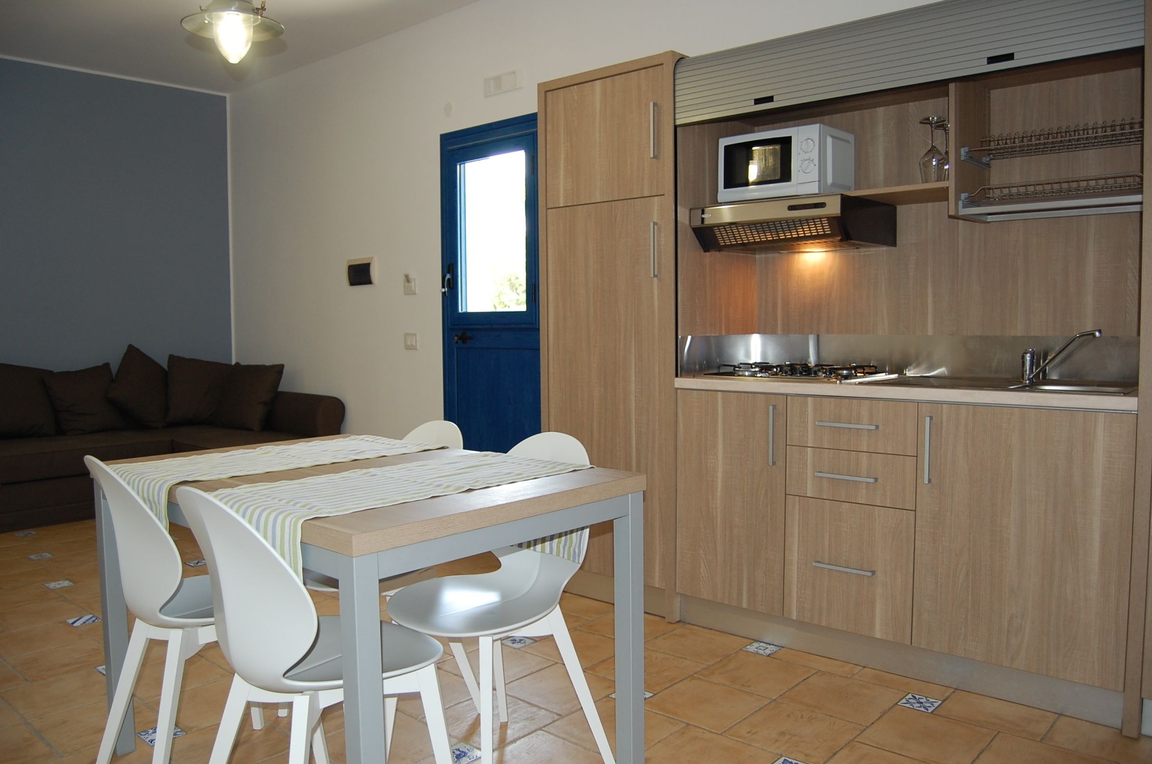 A compact kitchen with wooden cabinets, a microwave, and a sink. There is a dining area with a table and chairs.