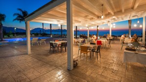 An outdoor dining area with tables and chairs, a pool, and a view of the sea and mountains.