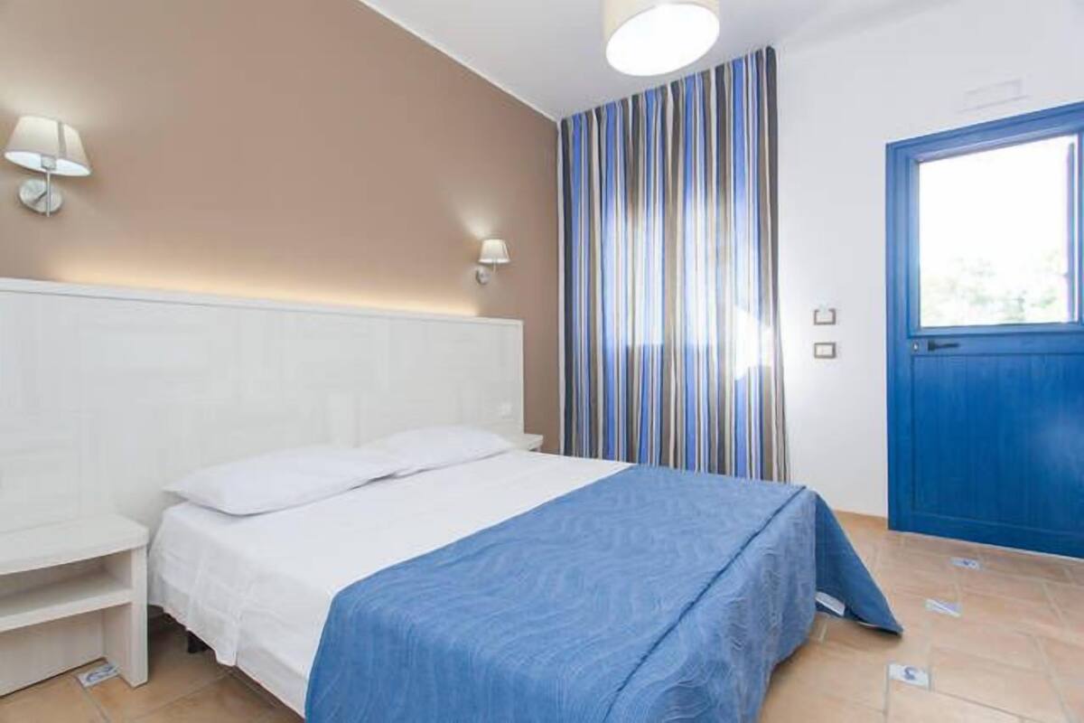 A bedroom with a bed, a nightstand, a lamp, a window with curtains, and a door.