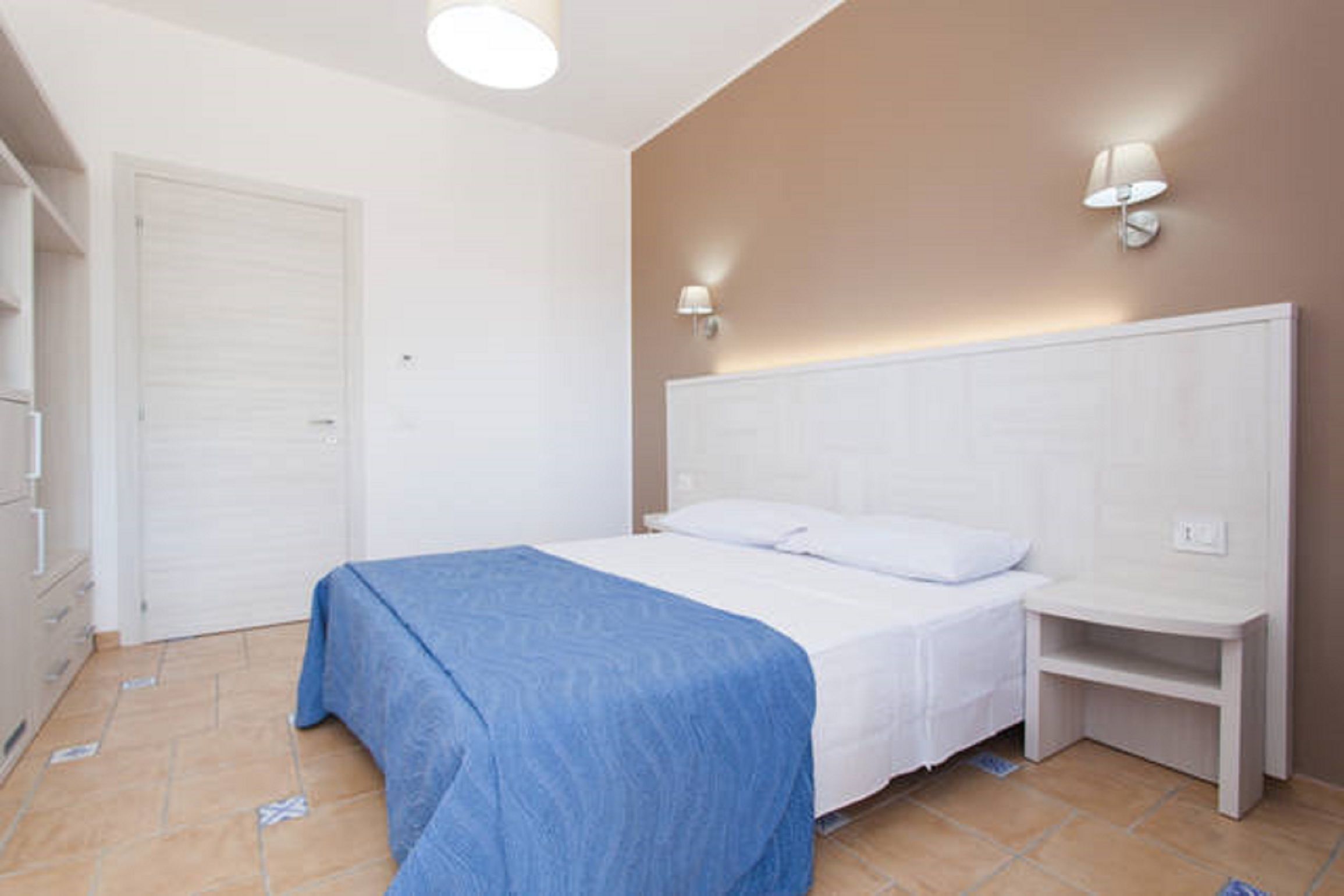 Une chambre à coucher avec un lit, une table de chevet, une lampe fixée au mur et une porte.