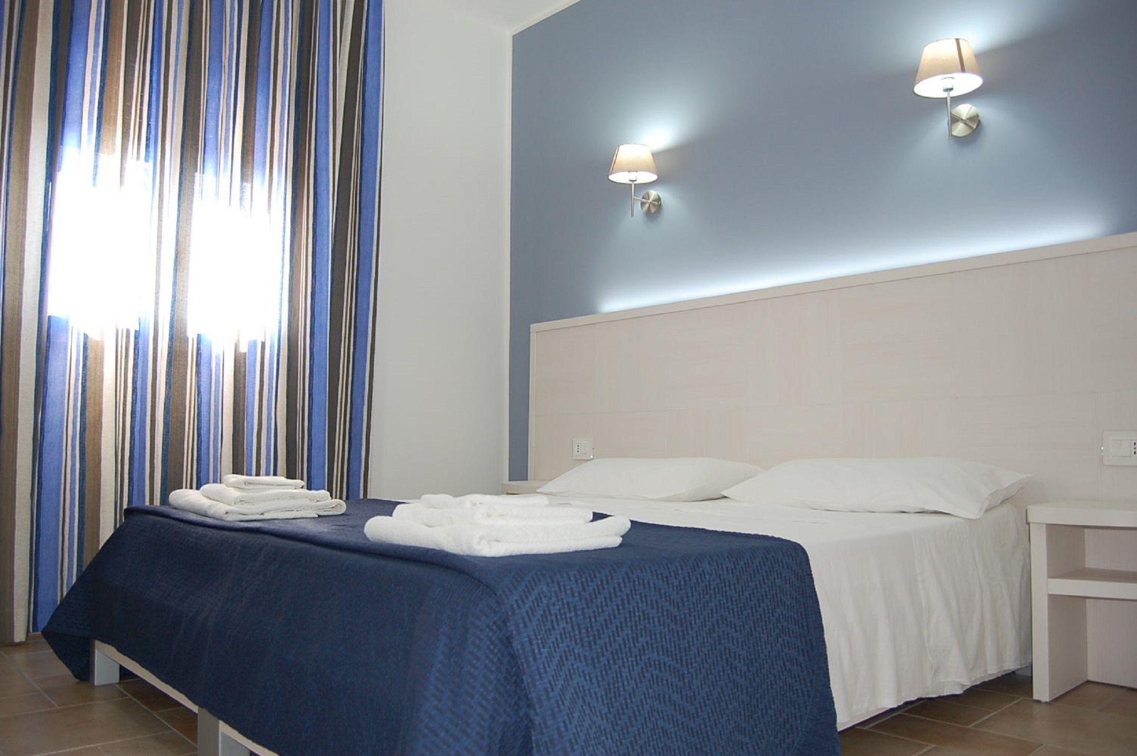 Ein Hotelzimmer mit einem Bett, weißen Kissen, einer blauen Bettdecke, zwei Wandleuchten und einem Fenster mit vertikalen Rollläden.