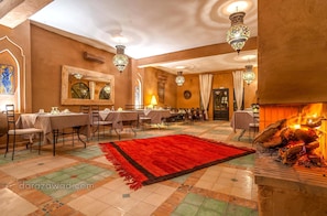 Un área de comedor con mesas dispuestas para huéspedes, un tapete rojo grande y una chimenea con leña encendida.
