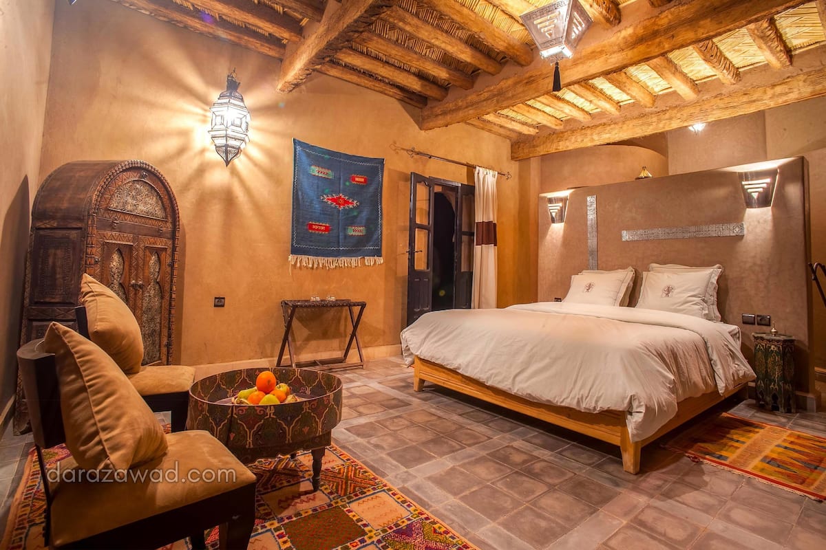 A traditional room with a bed, a chair, a small table with fruit, and a decorative wall hanging.