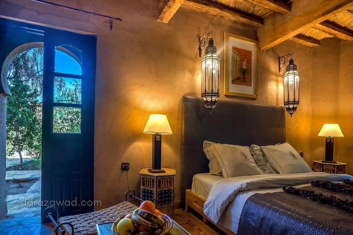 A cozy bedroom with a bed, two wall-mounted lamps, a small table with a lamp, and a door leading outside.