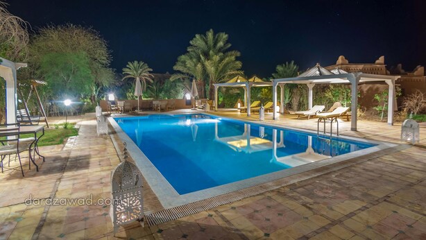 Una piscina con sillas de descanso y una pérgola, rodeada de palmeras y un cielo nocturno.