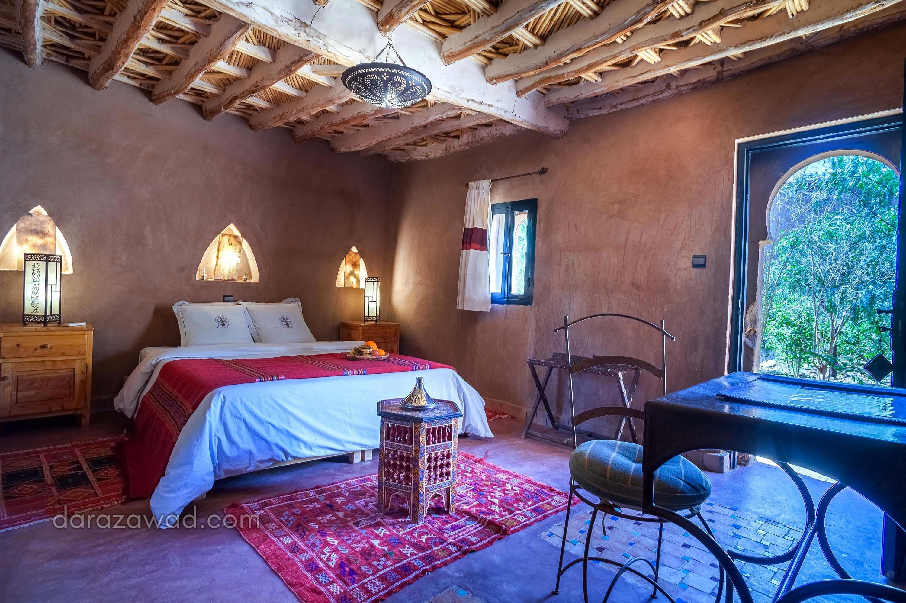 A bedroom with a wooden ceiling, a bed with a red blanket, two bedside lamps, a small table with a lantern, a chair, and a window with a view of greenery.