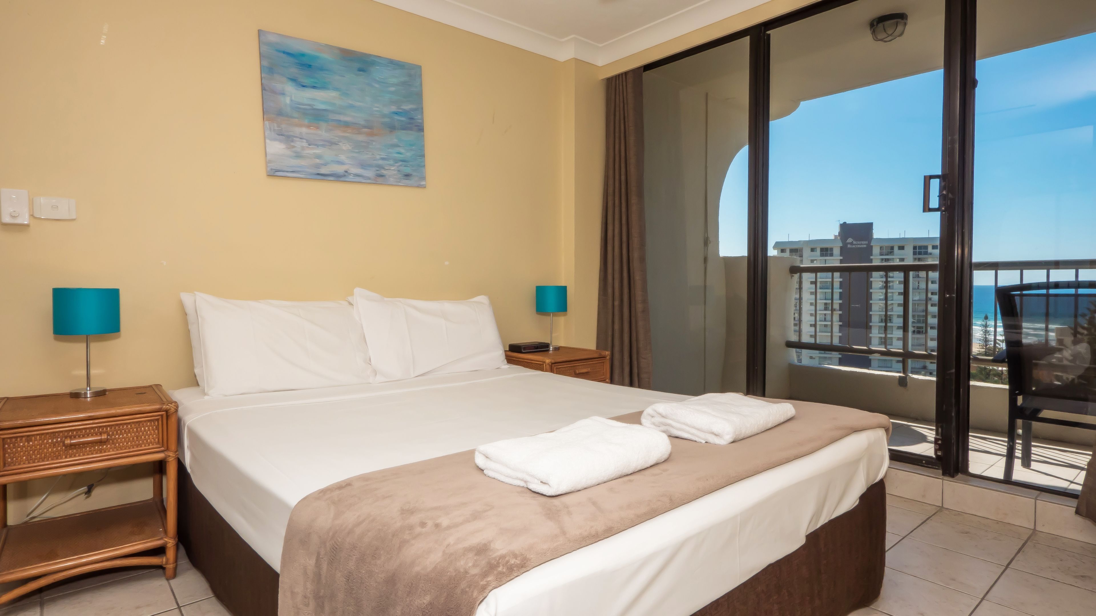 Una camera d'hotel con un letto, due comodini con lampade, un quadro appeso e un balcone vista su edifici e mare.