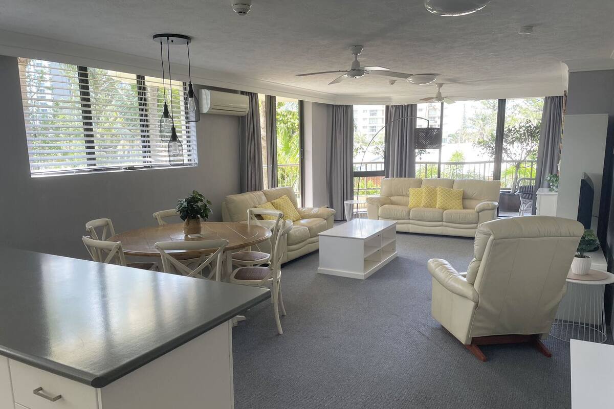 A modern living room with a dining area, large windows, ceiling fans, and a flat-screen TV.